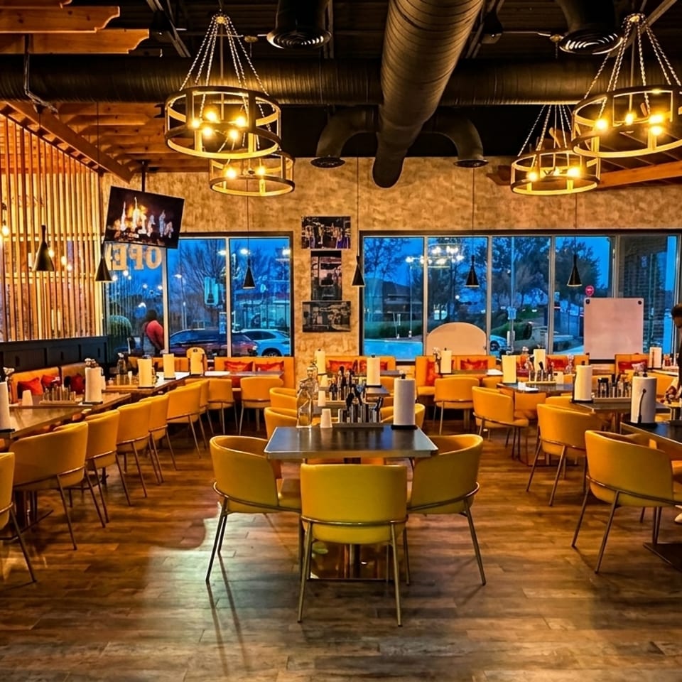 Spacious restaurant interior with yellow chairs, dark tables, warm lighting, and two staff members preparing drinks.